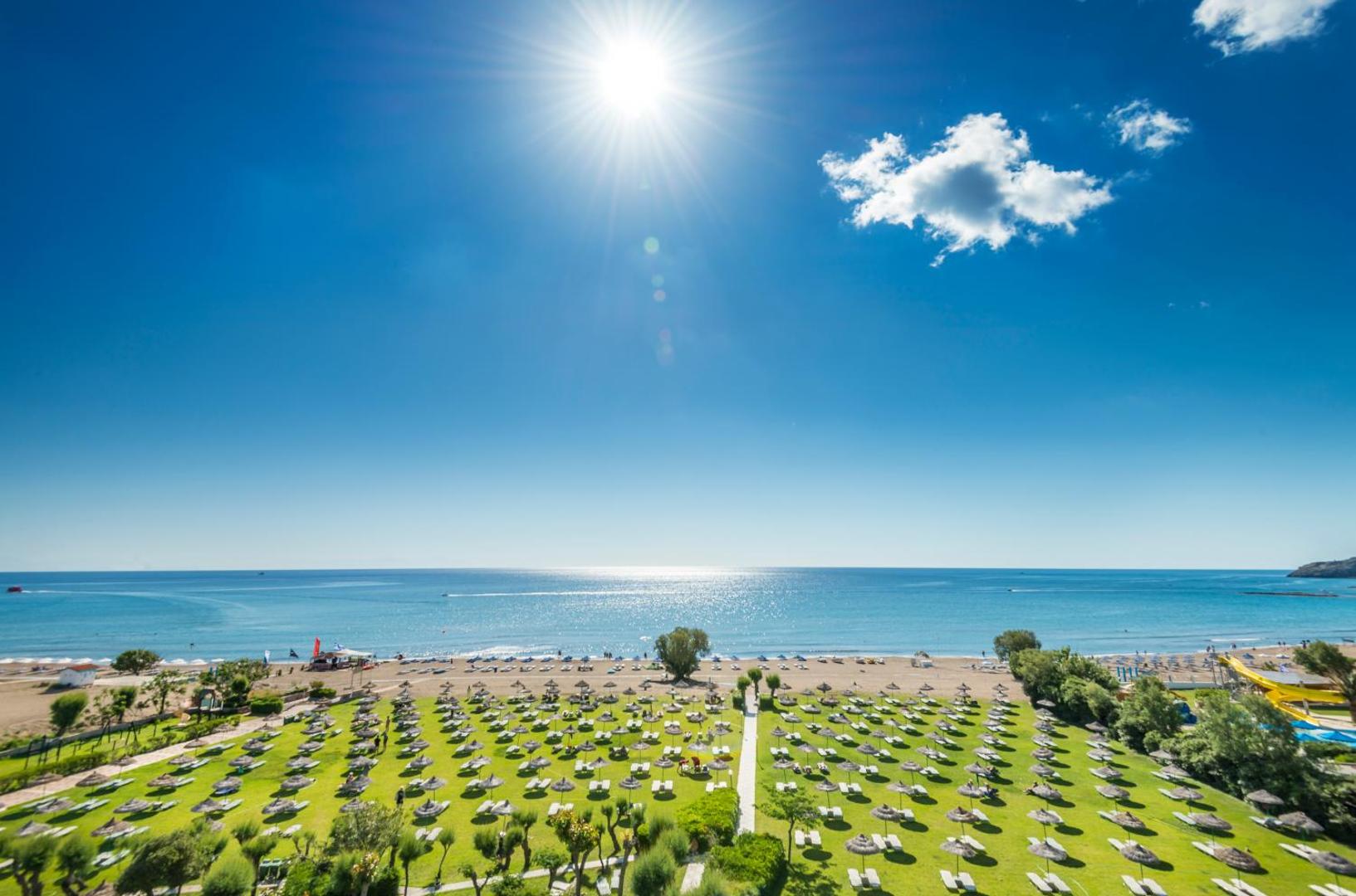 Panorāmas skats uz Apollo Beach teritoriju Rodā — zaļš zāliens ar saulessargiem, smilšu pludmale un dzidra jūra saulainā dienā