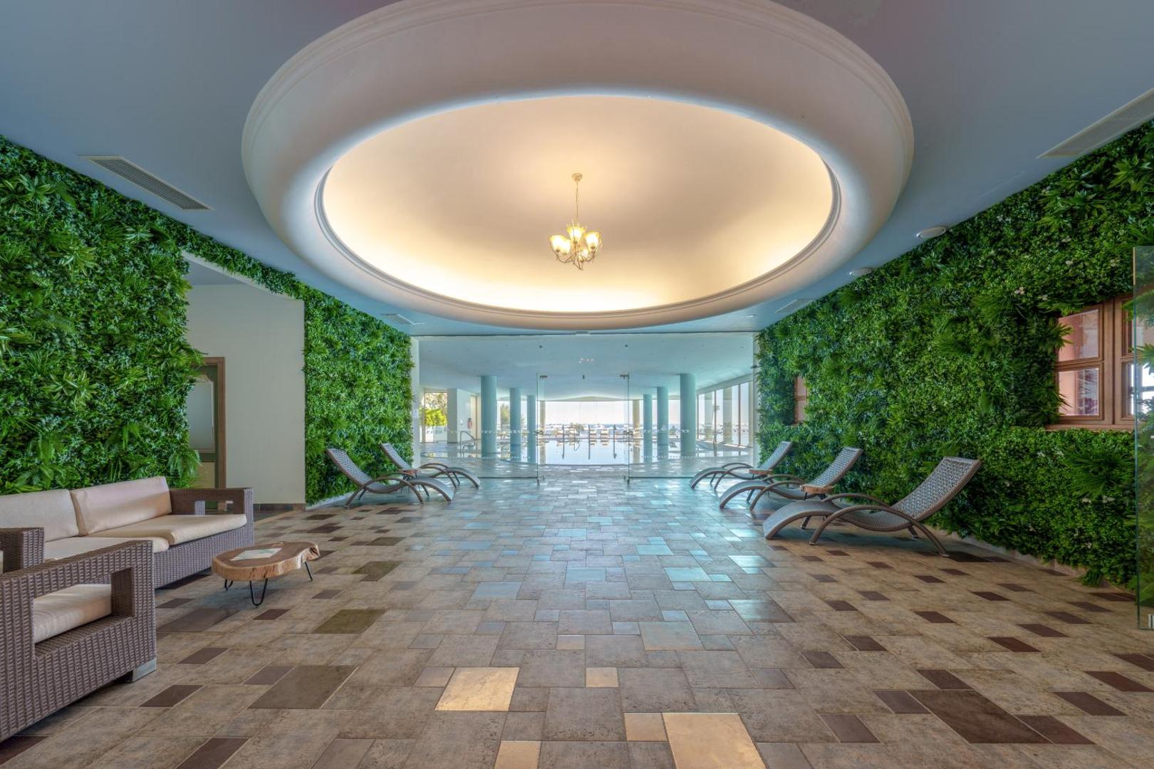 Atrium Prestige viesnīcas spa atpūtas zona ar zaļajām sienām, ērtiem zviļņiem un skatu uz iekštelpu baseinu Rodā