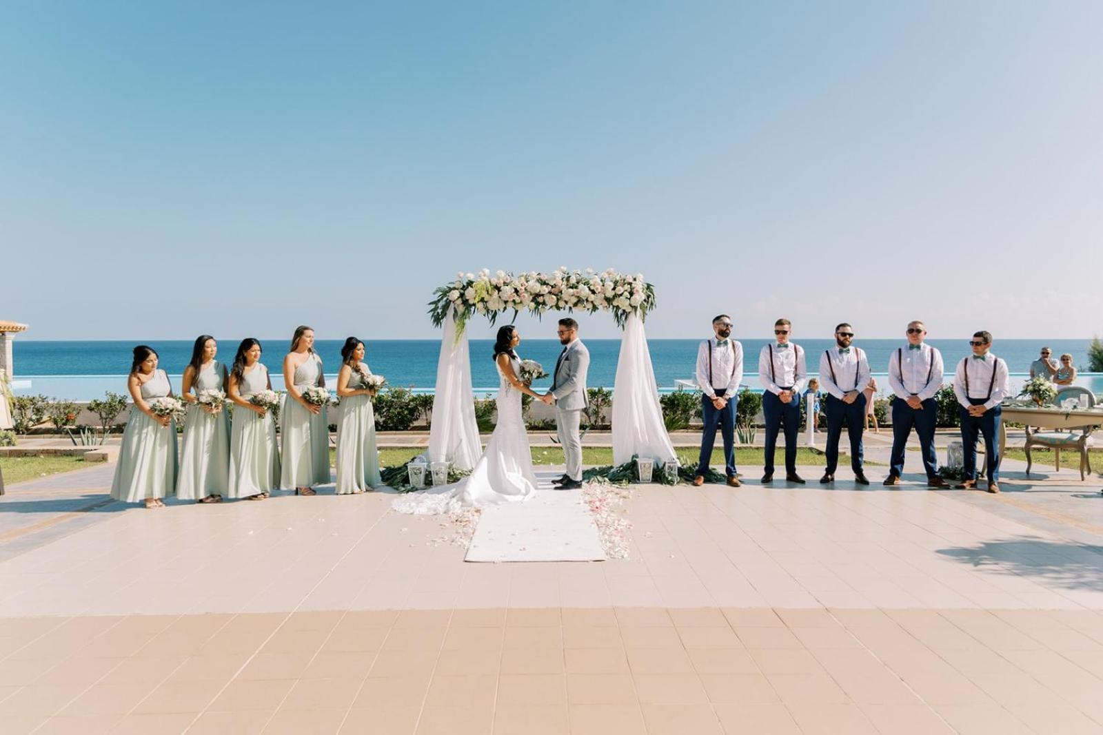 Kāzu ceremonija Atrium Prestige Thalasso Spa Resort & Villas terasē pie jūras ar ziedu arku, jauno pāri un kāzu viesiem Rodā.