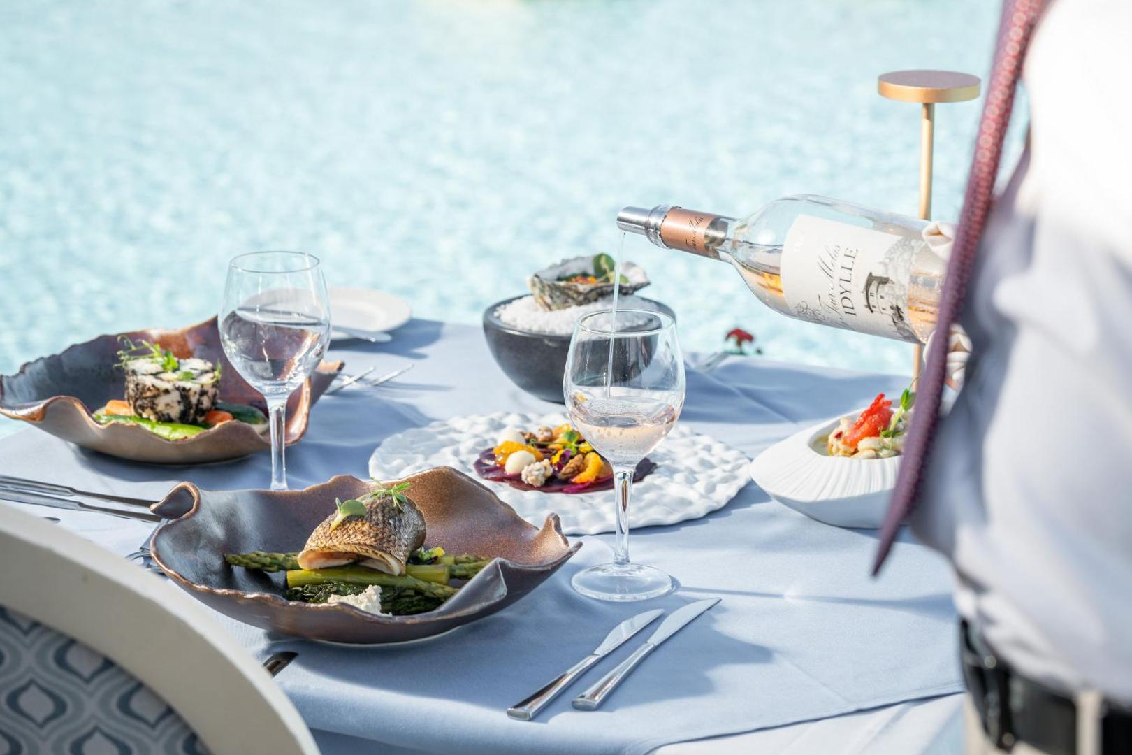 Viesmīlis lej vīnu glāzē pie restorāna galdiņa Atrium Prestige Thalasso Spa Resort piekrastē, fonā redzams baseins un Vidusjūras ainava.