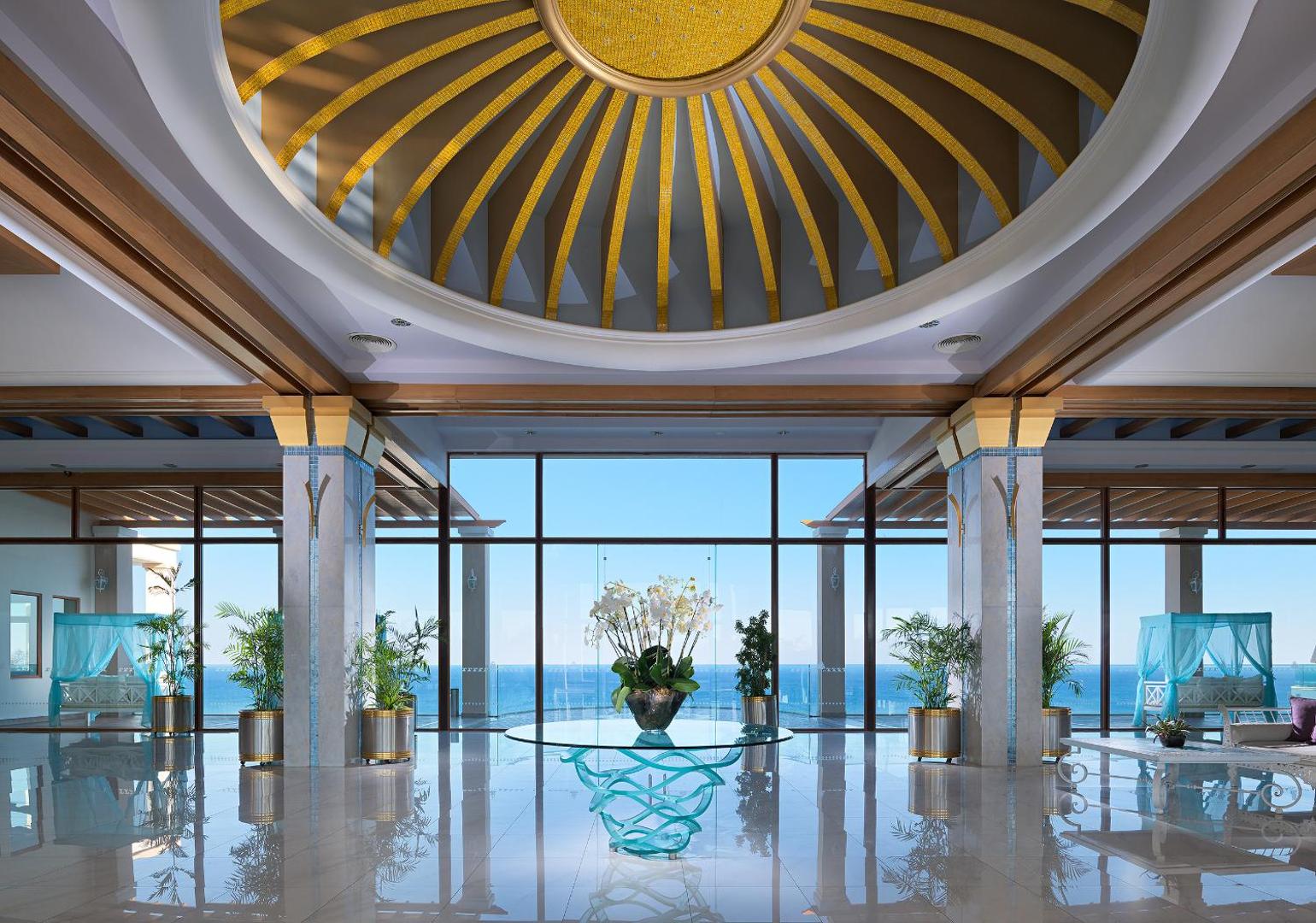 Atrium Prestige viesnīcas lobby ar greznu kupolu un panorāmas logiem uz jūru Rodas salā — plaša un gaiša uzņemšanas zāle