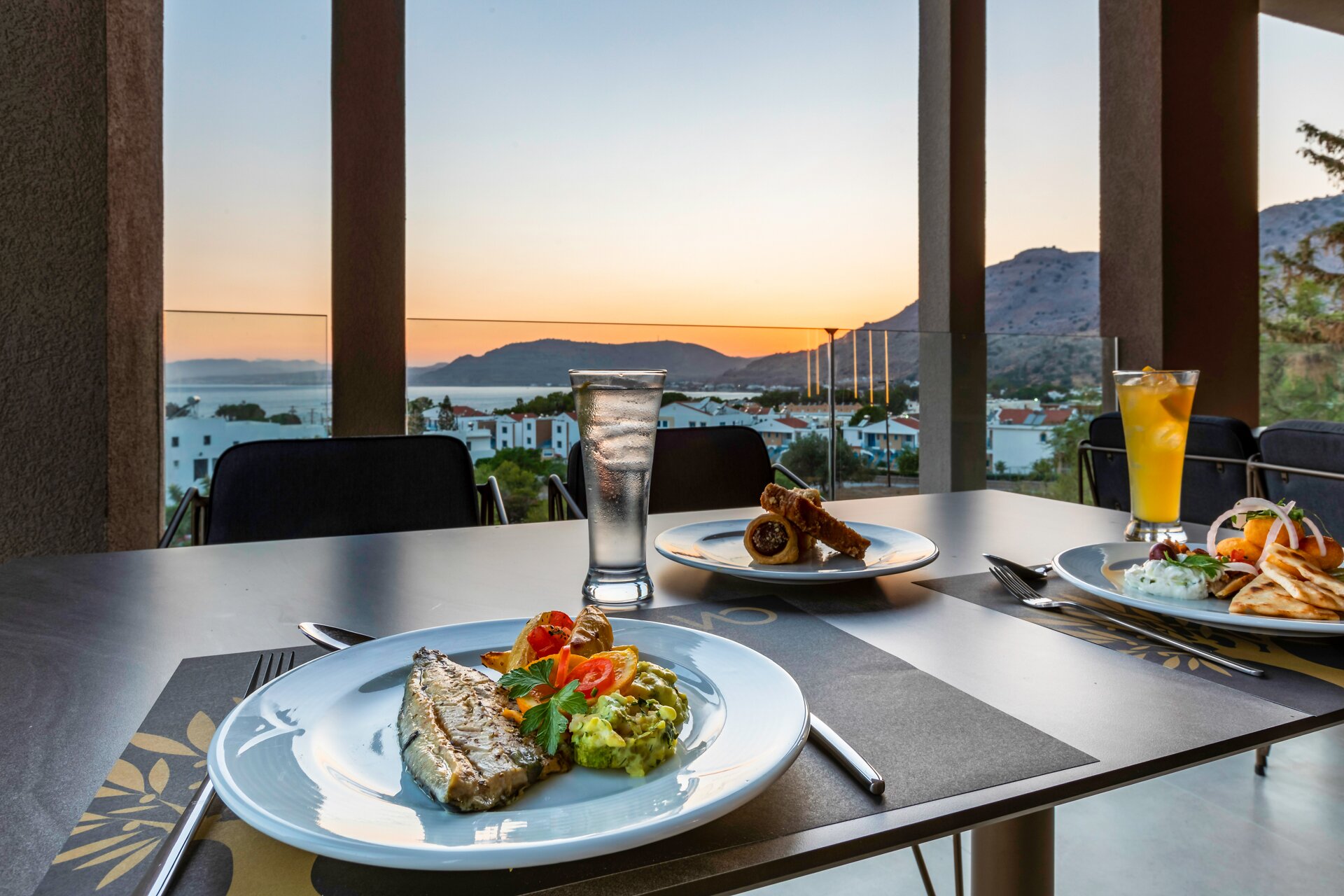 Viesnīcas White Olive Premium Lindos restorāna terase ar klātu galdu, zivju ēdienu un panorāmas skatu uz kalniem un jūru