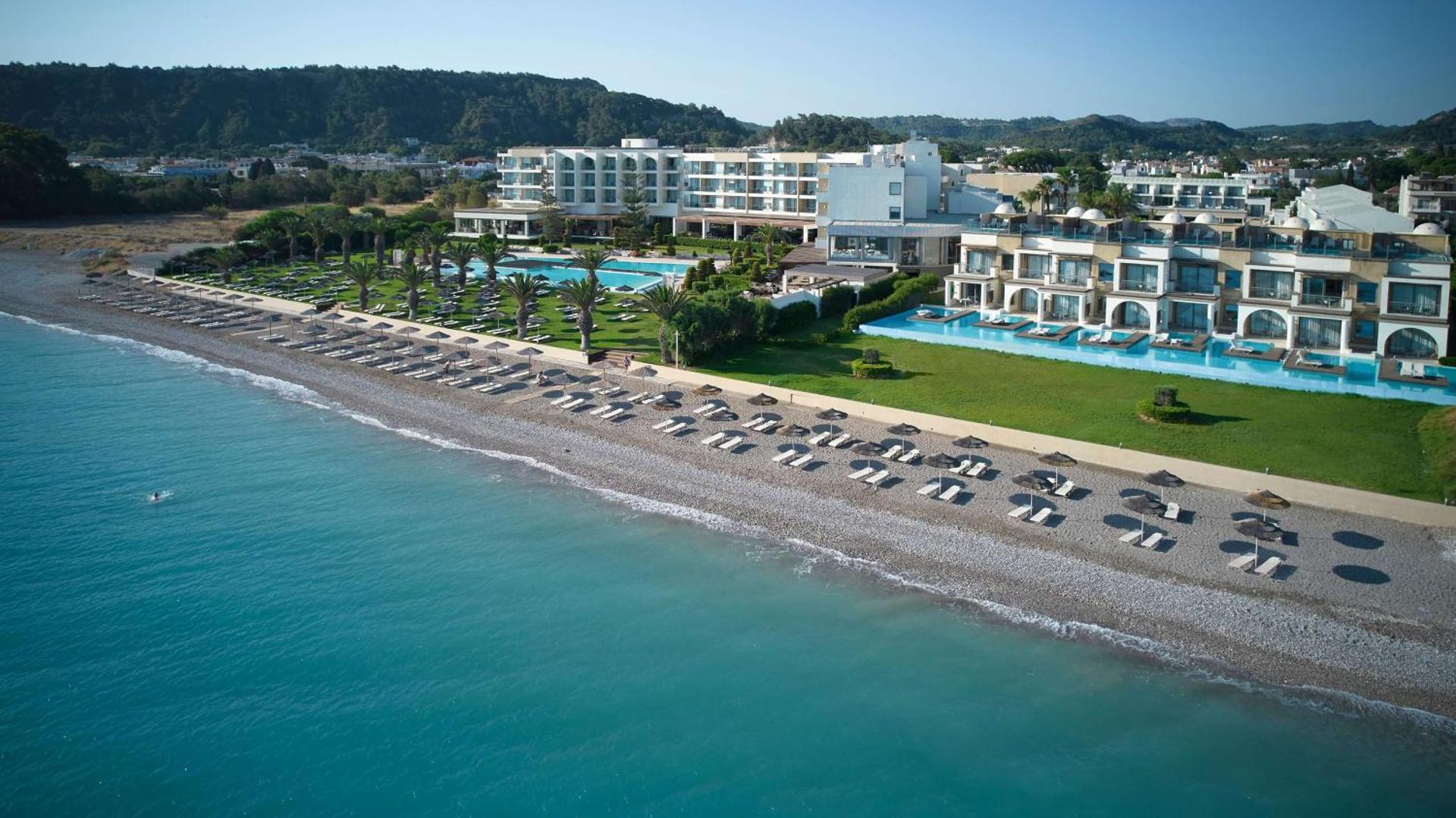 The Ixian Grand & All Suites viesnīcas panorāmas skats Rodas piekrastē ar plašu oļu pludmali, baseiniem un modernu arhitektūru.