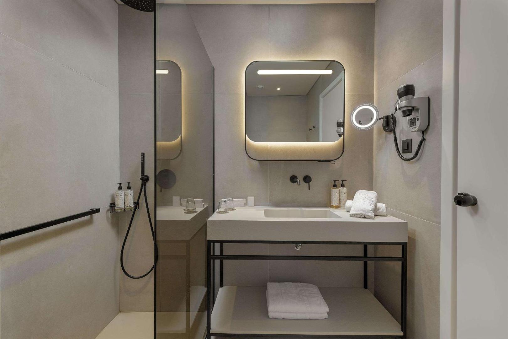 The Ixian Grand & All Suites modernā numura vannasistaba ar izgaismotu spoguli, dušu un gaišām flīzēm Rodas kūrortā