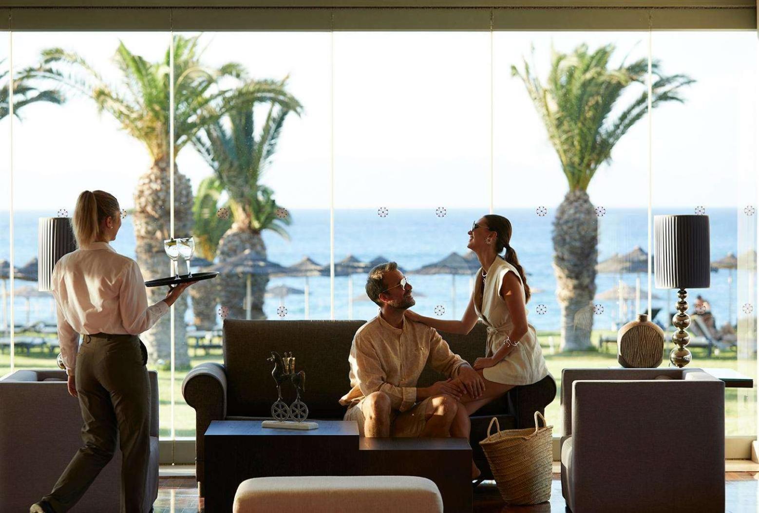 Viesi bauda dzērienus The Ixian Grand & All Suites lobby bārā ar lieliem panorāmas logiem uz dārzu un jūru
