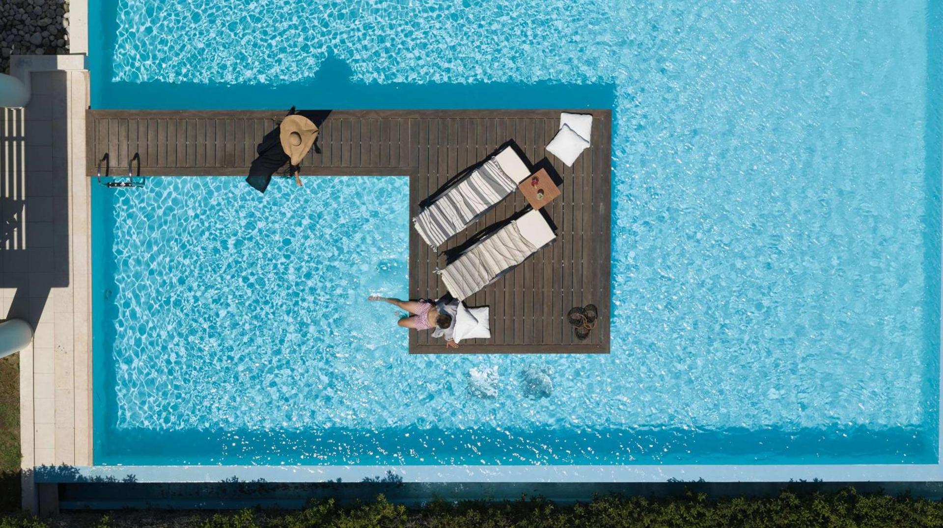 The Ixian Grand & All Suites skats no augšas uz baseinu ar koka terasi, sauļošanās krēsliem un atpūtniekiem Rodas salā