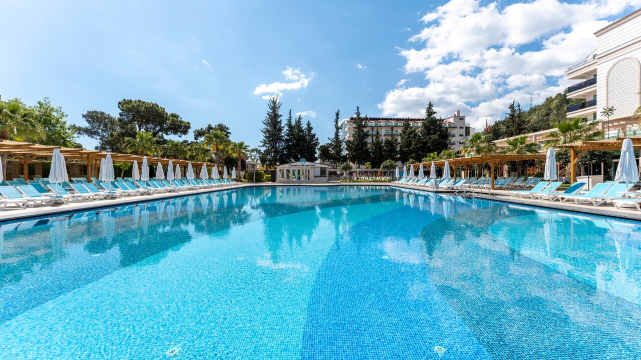 Plašs Dosinia Luxury Resort peldbaseins ar dzidru ūdeni, apkārt izvietotiem saulessargiem un palmām saulainā dienā