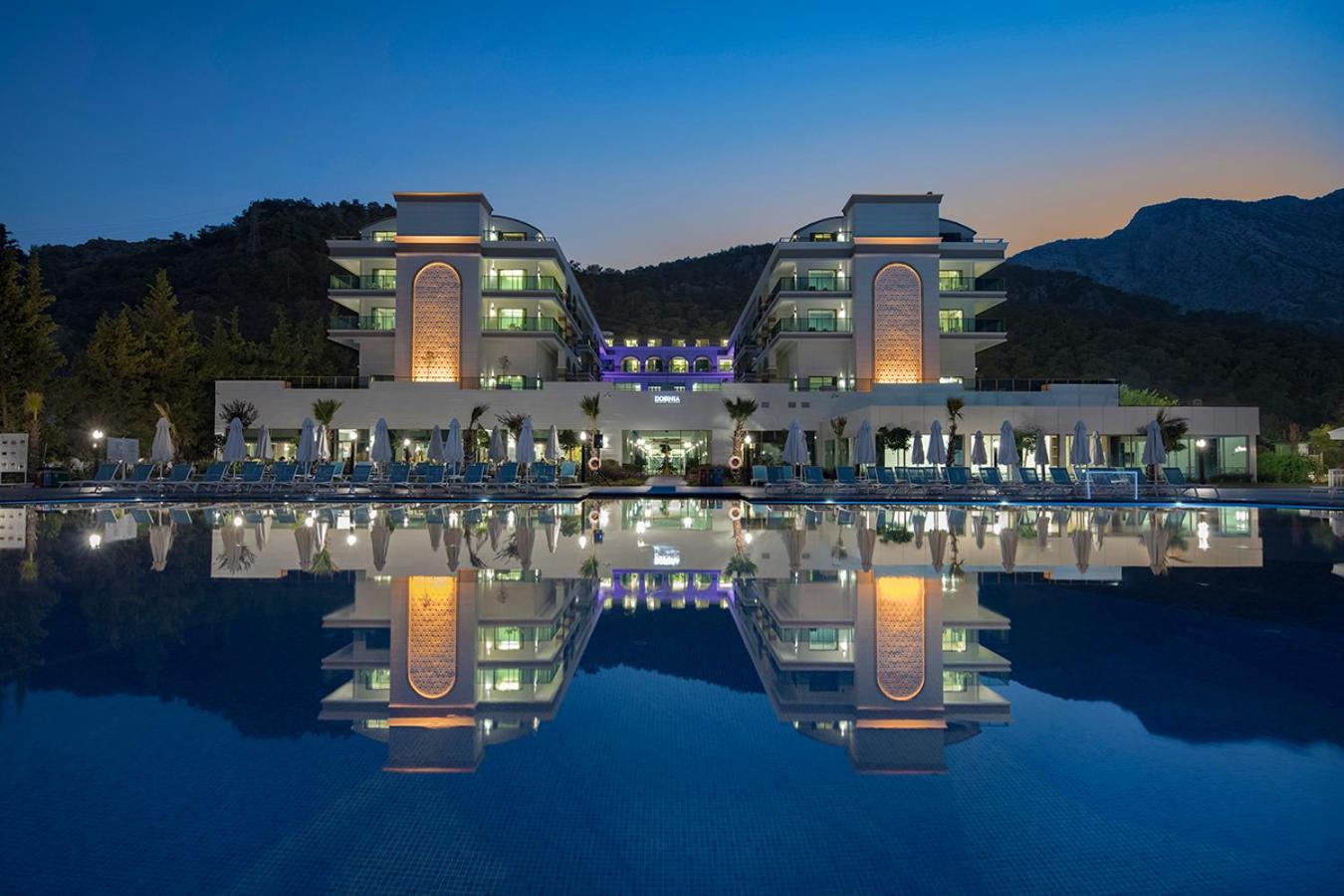 Dosinia Luxury Resort fasāde un baseins vakarā ar izgaismotiem arhitektūras elementiem un atspulgu ūdenī Beldibi reģionā