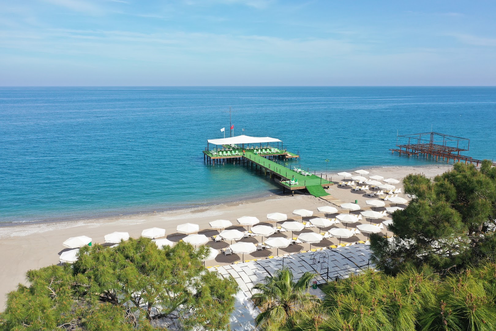 Dosinia Luxury Resort pludmales zona ar baltiem saulessargiem un zaļu koka laipu, kas ved uz piestātni Vidusjūras krastā