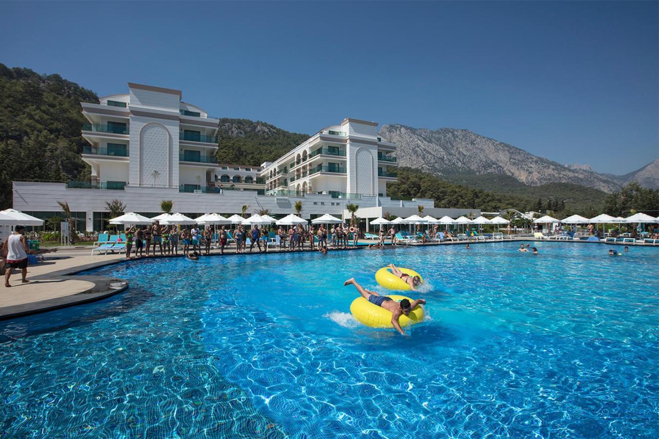 Dosinia Luxury Resort āra baseins ar atpūtniekiem uz dzelteniem piepūšamajiem riņķiem, fonā viesnīcas ēka un kalni Kemeras reģionā