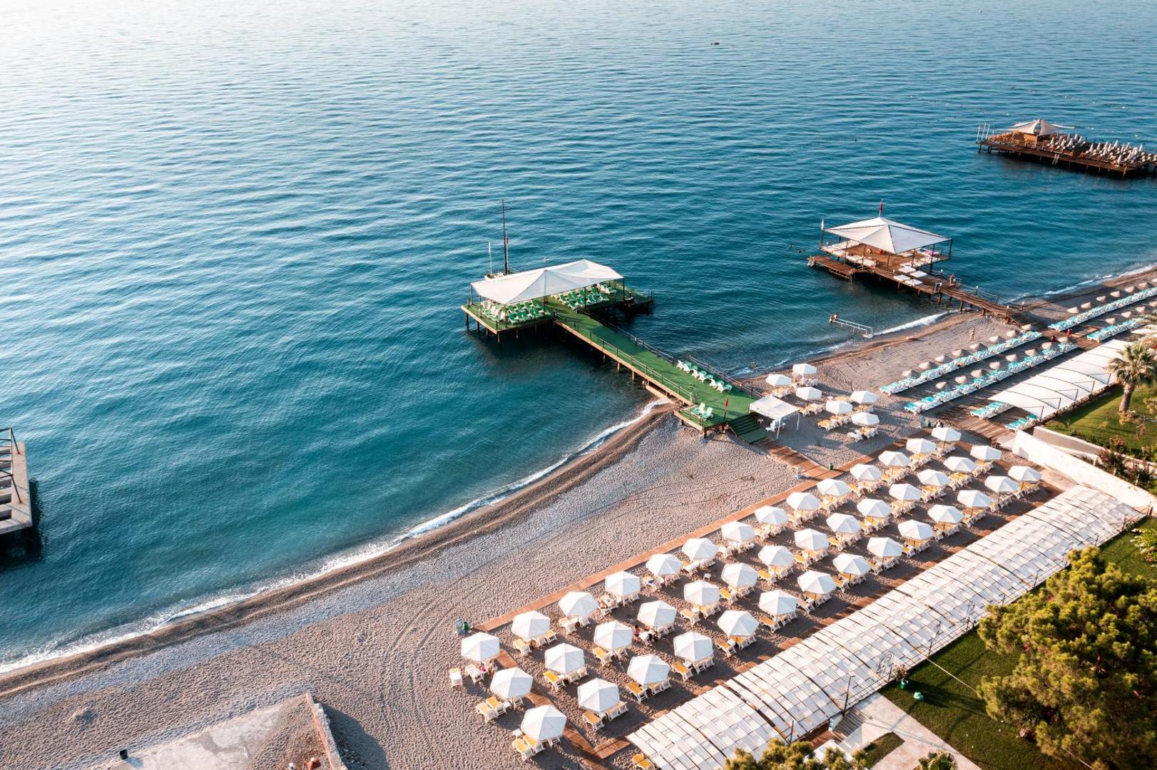 Skats no augšas uz Dosinia Luxury Resort oļu pludmali, saulessargu rindām un divām koka piestātnēm saulainā dienā Turcijā