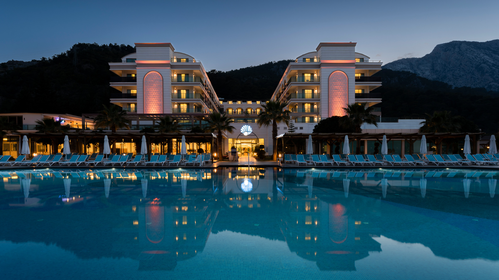 Dosinia Luxury Resort fasāde un izgaismots baseins vakarā, atspulgs ūdenī un kalnu siluets fonā Kemeras piekrastē