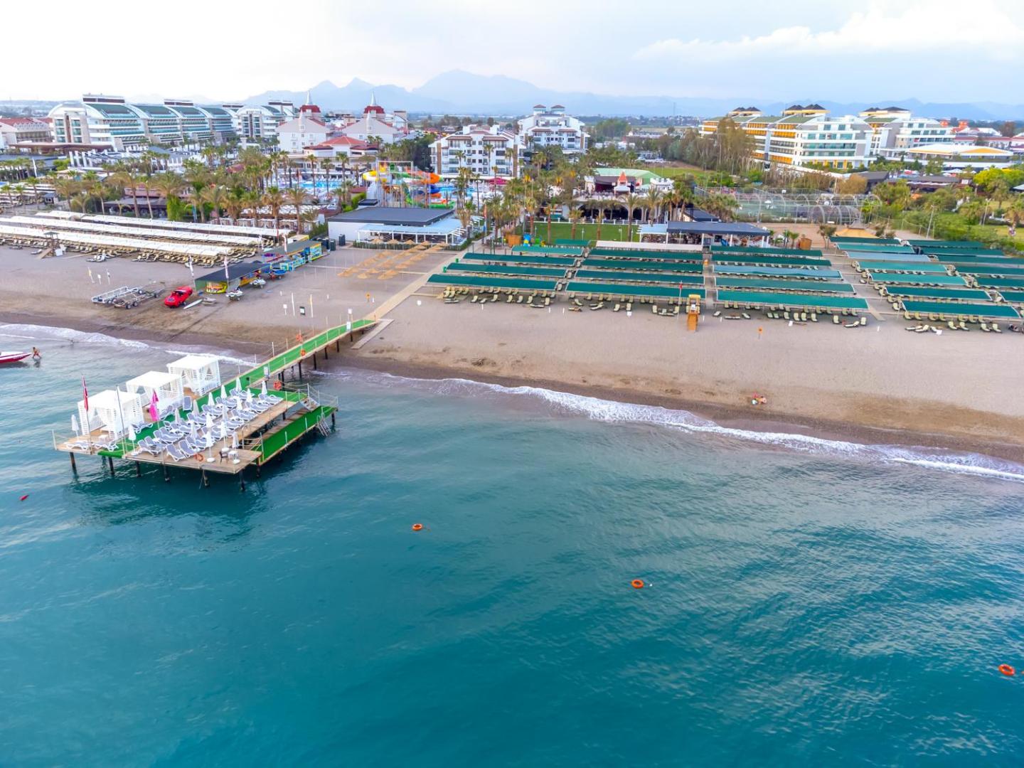 Belek Beach Resort viesnīcas panorāmas skats no putna lidojuma uz Vidusjūras piekrasti, privāto pludmali, piestātni un plašo baseinu zonu Turcijā.
