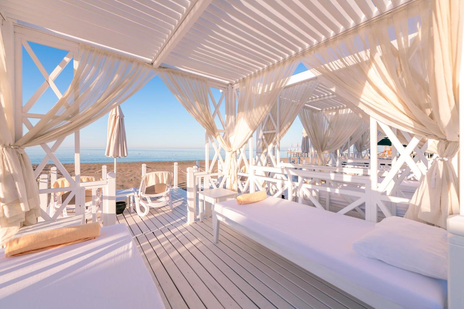 Belek Beach Resort pludmales atpūtas zonas balti koka bungalo ar aizkariem un mīkstiem matračiem tiešā jūras tuvumā saulainā dienā.