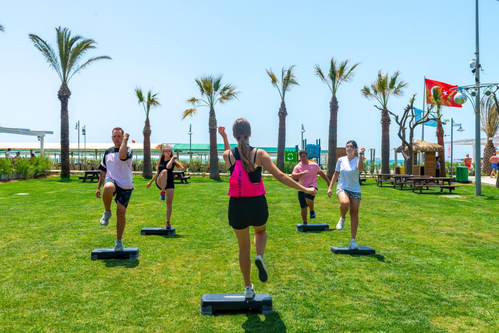 Viesnīcas Belek Beach Resort viesi piedalās rīta aerobikas nodarbībā uz zaļā zālāja palmu ieskautā teritorijā pie jūras.