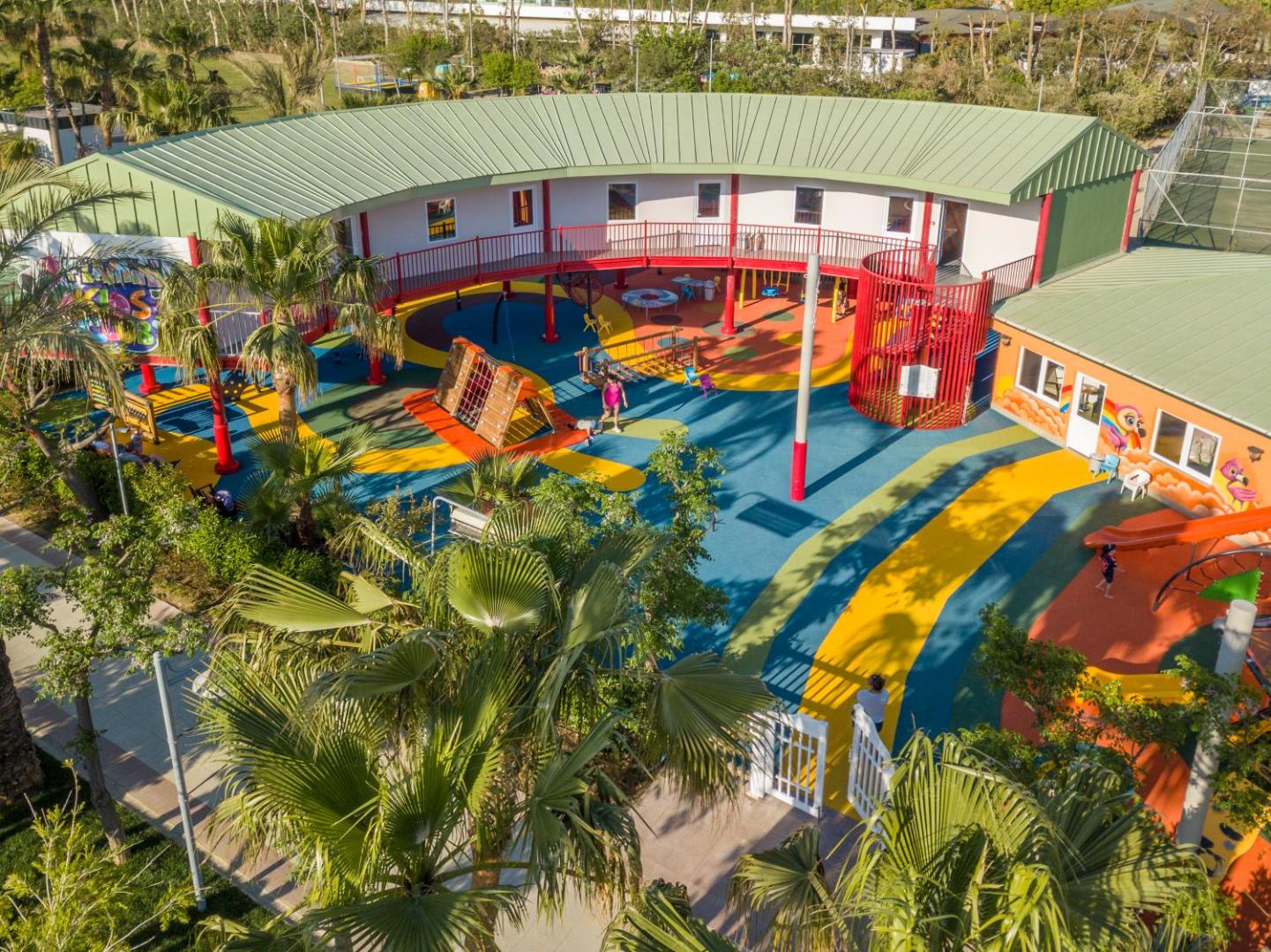 Belek Beach Resort bērnu kluba āra rotaļu laukums no augšas — krāsains segums, kāpšļi un palmas viesnīcas dārzā Belekā.