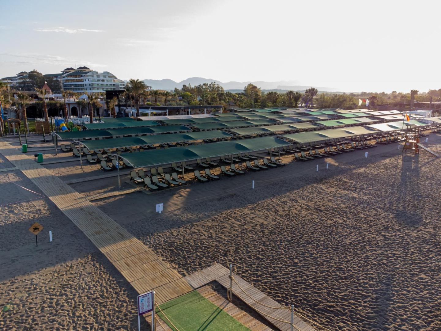 Belek Beach Resort plašā smilšu pludmale ar daudzām sauļošanās krēslu rindām zem kopējām nojumēm un koka laipām piekrastes zonā.