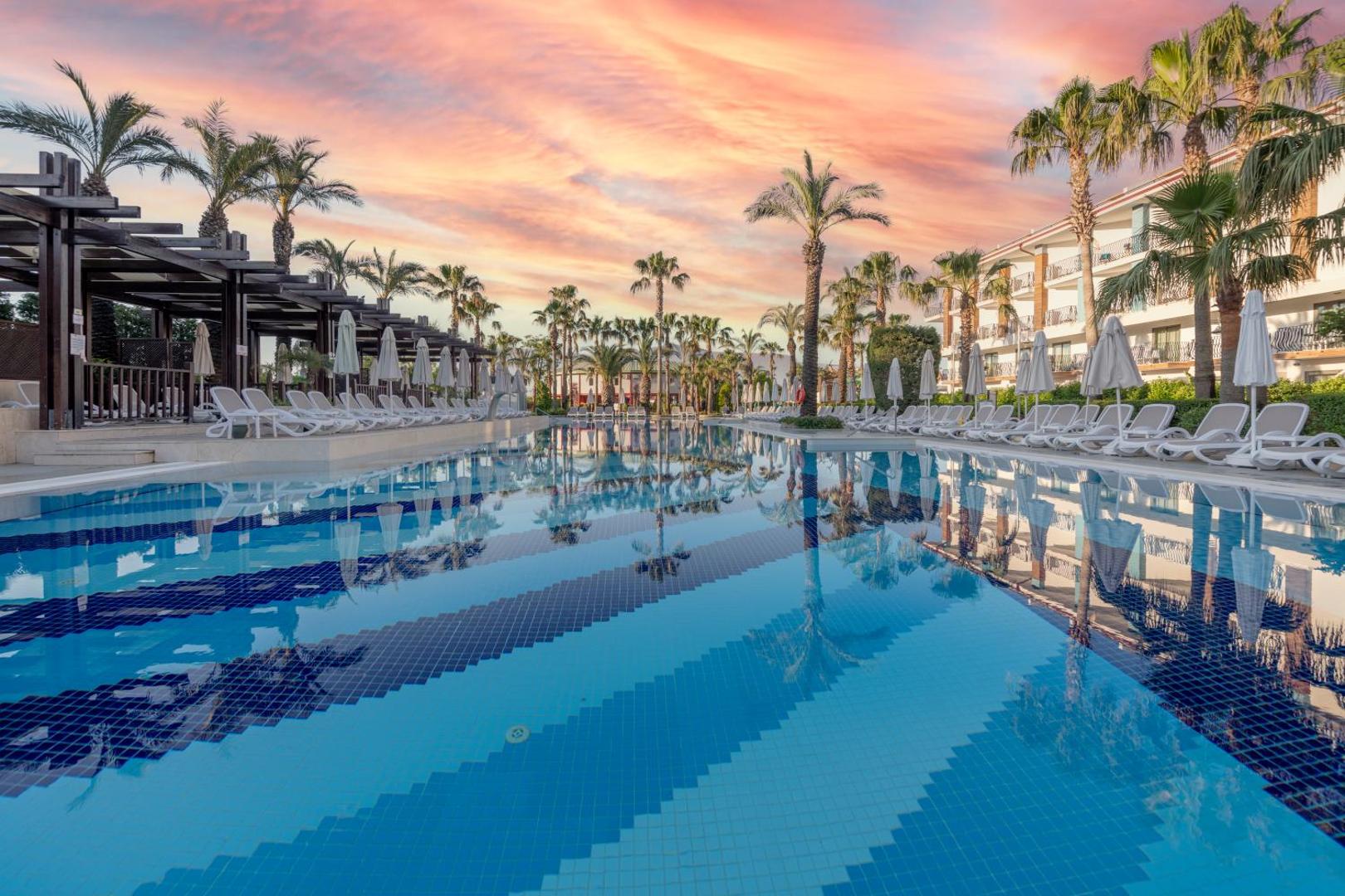 Belek Beach Resort lielais āra baseins saulrieta laikā ar palmām, sauļošanās krēsliem un viesnīcas ēkām fonā