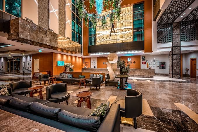 Belek Beach Resort modernais viesnīcas lobby ar augstiem griestiem, dizaina lustrām, marmora grīdām un ērtām atpūtas mēbelēm recepcijas zonā.