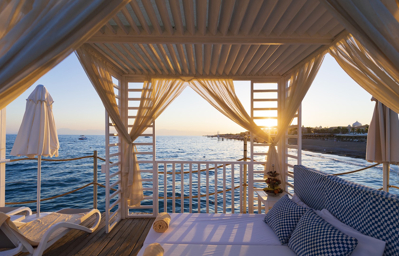 Saulrieta skats no Belek Beach Resort piestātnes bungalo uz Vidusjūru, mīkstie spilveni un augļu plate romantiskai atmosfērai vakarā.