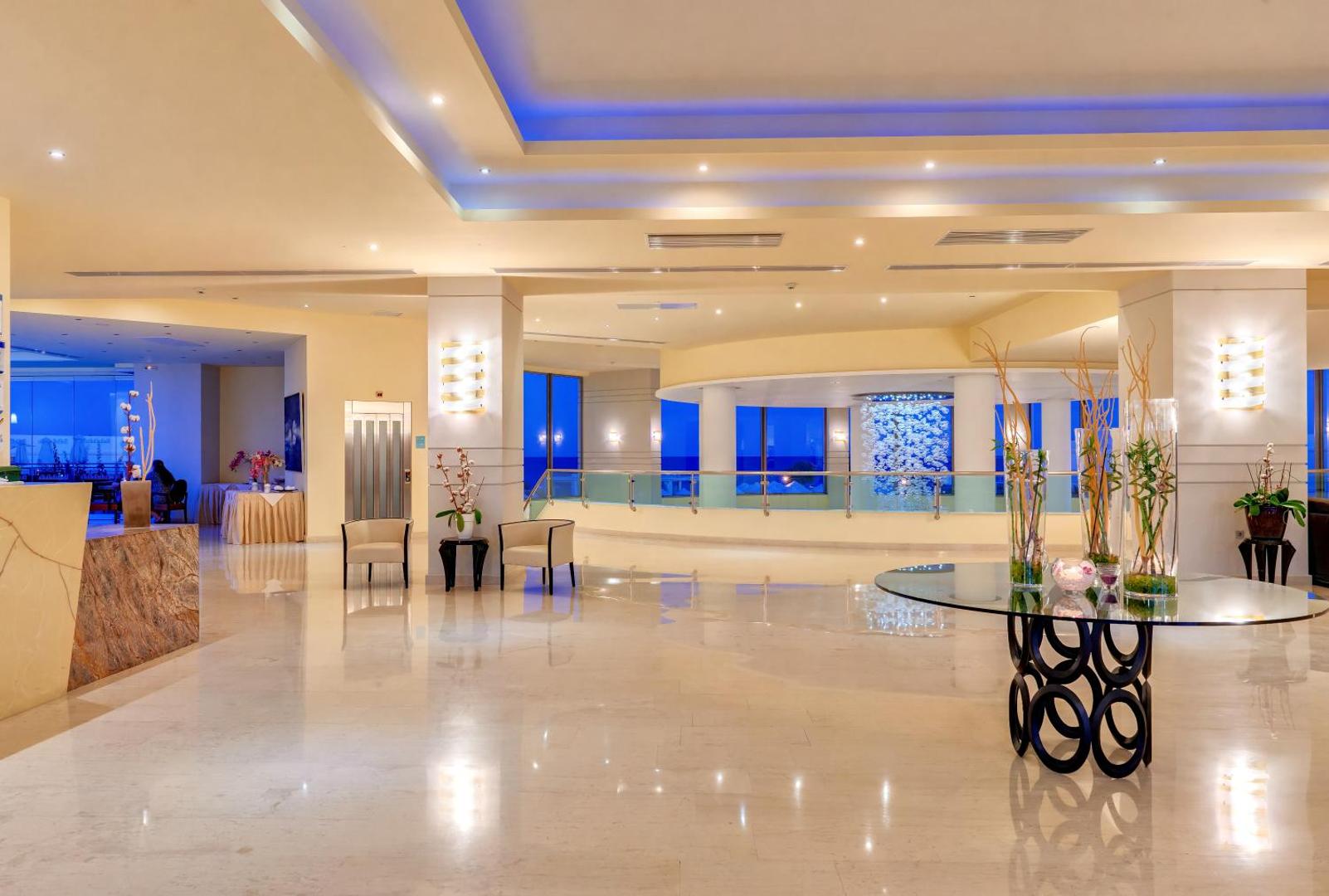 Apollo Blue Hotel modernais lobby interjers ar dizaina elementiem, stikla galdiem un skatu uz vakara gaismām caur lielajiem logiem.