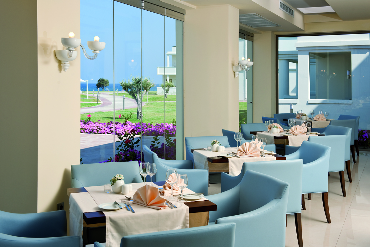 Apollo Blue Hotel gaišā restorāna zāle ar panorāmas logiem, skatu uz dārzu un jūru, klātiem galdiem un ziliem krēsliem Faliraki.