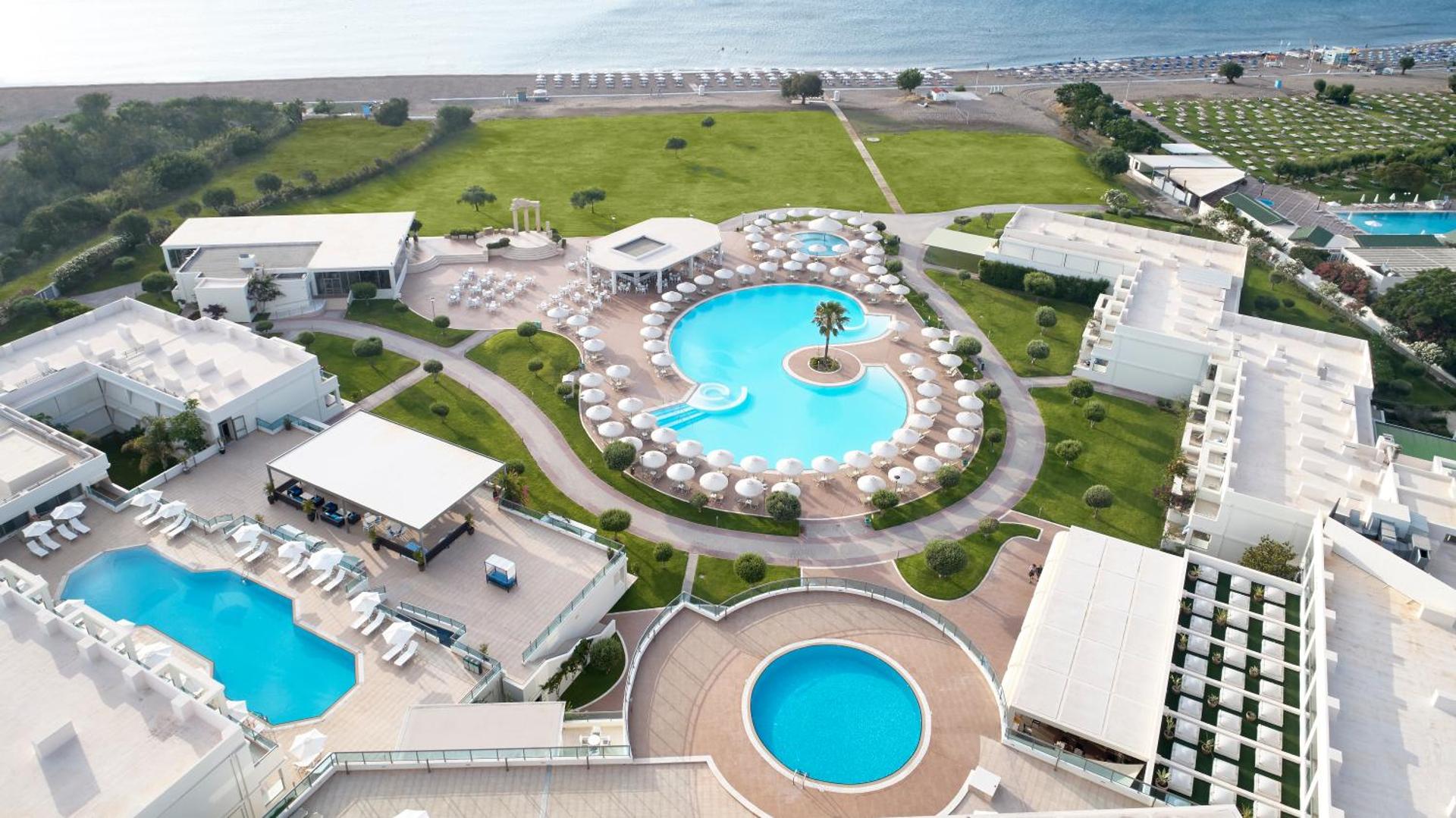 Apollo Blue Hotel teritorijas skats no putna lidojuma Rodas salā — baseinu komplekss, zaļie zālāji un tieša piekļuve oļu pludmalei
