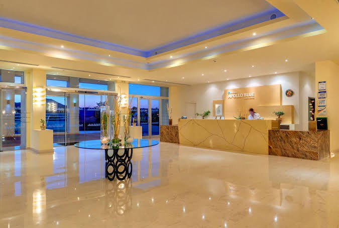 Apollo Blue Hotel plašais un modernais lobby vestibils ar marmora grīdu, recepcijas leti un stilīgu apgaismojumu Rodas salā, Grieķijā.