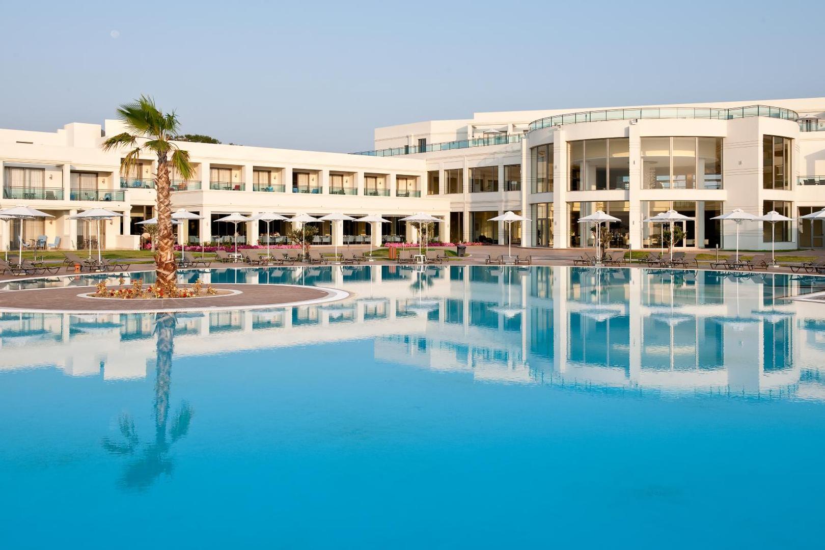 Apollo Blue Hotel galvenais baseins ar palmu un viesnīcas ēkas atspulgu ūdenī — saulessargi un atpūtas krēsli Rodas piekrastē