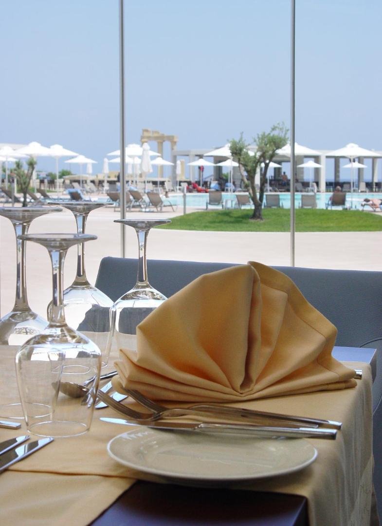 Skats no Apollo Blue Hotel restorāna galda uz āra baseinu un saulessargiem caur stikla sienu, elegants galda klājums priekšplānā.