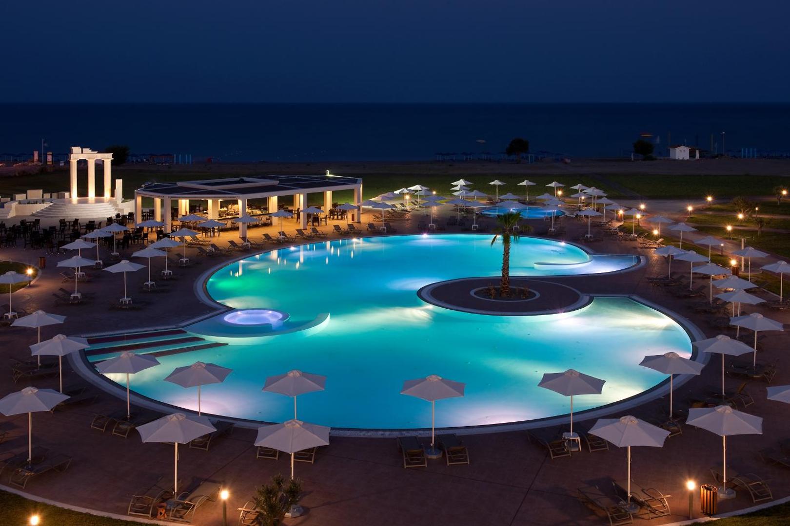 Apollo Blue Hotel āra baseins vakarā ar izgaismotu bāru un dekoratīvām kolonnām — romantiska atmosfēra Rodas salas piekrastē
