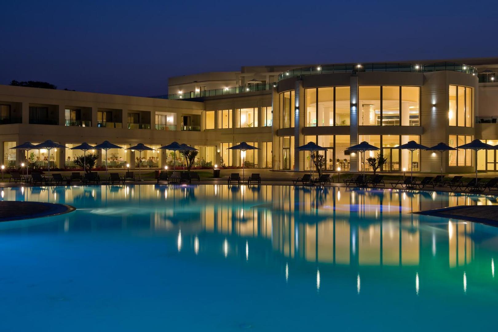 Apollo Blue Hotel vakara skats uz izgaismoto baseinu un restorāna zonu — lieli logi un gaismu atspulgi ūdens virsmā