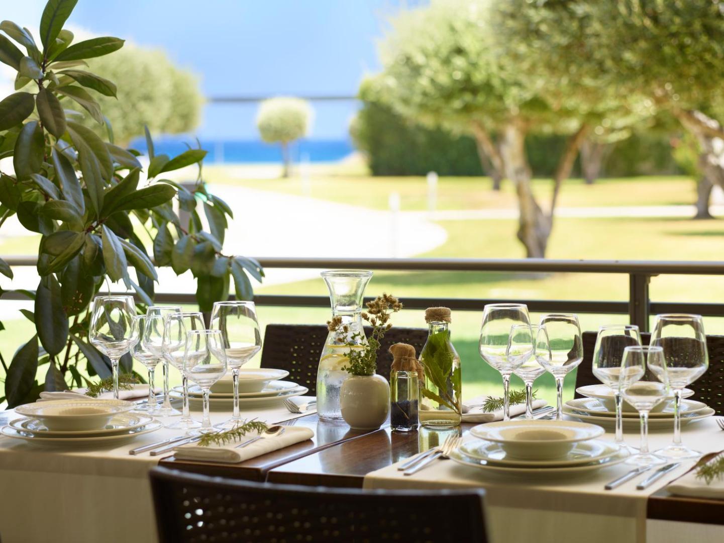 Apollo Blue Hotel restorāna terase ar eleganti klātu galdu, baltiem traukiem un skatu uz zaļo dārzu un jūru tālumā