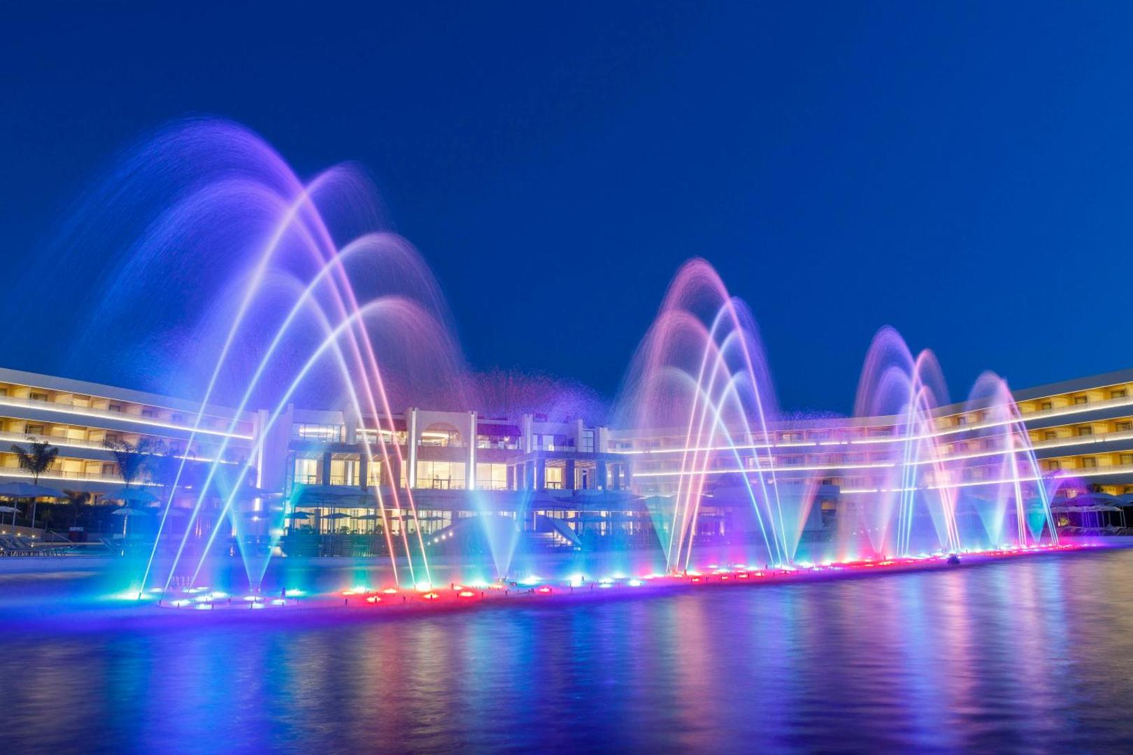 Princess Andriana Resort & Spa muzikālo strūklaku šovs ar krāsainu apgaismojumu uz viesnīcas ēku fona naktī