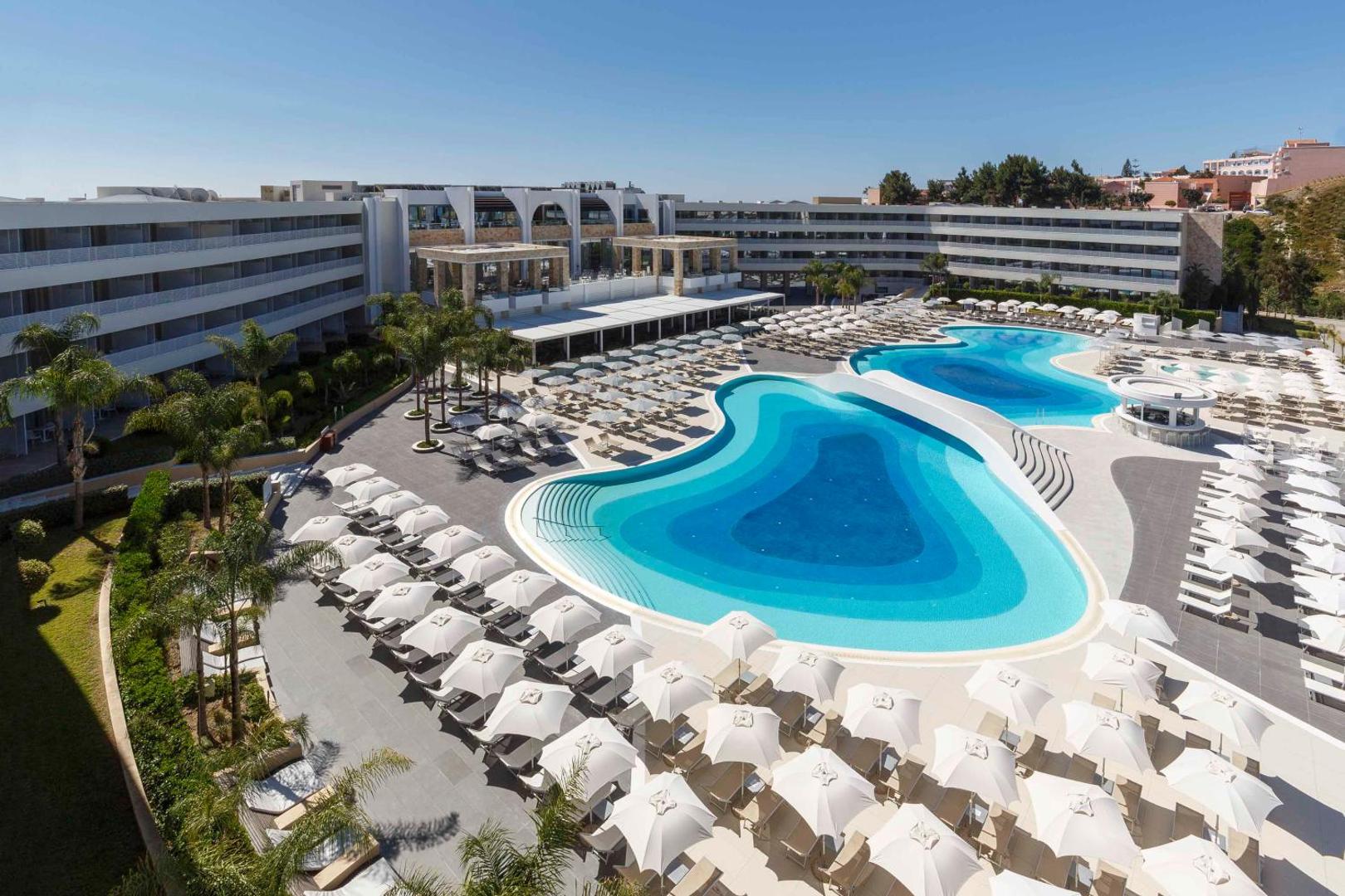 Princess Andriana Resort & Spa viesnīcas ēka un plašā baseina zona Rodā — balti saulessargi, sauļošanās krēsli un palmas piekrastē