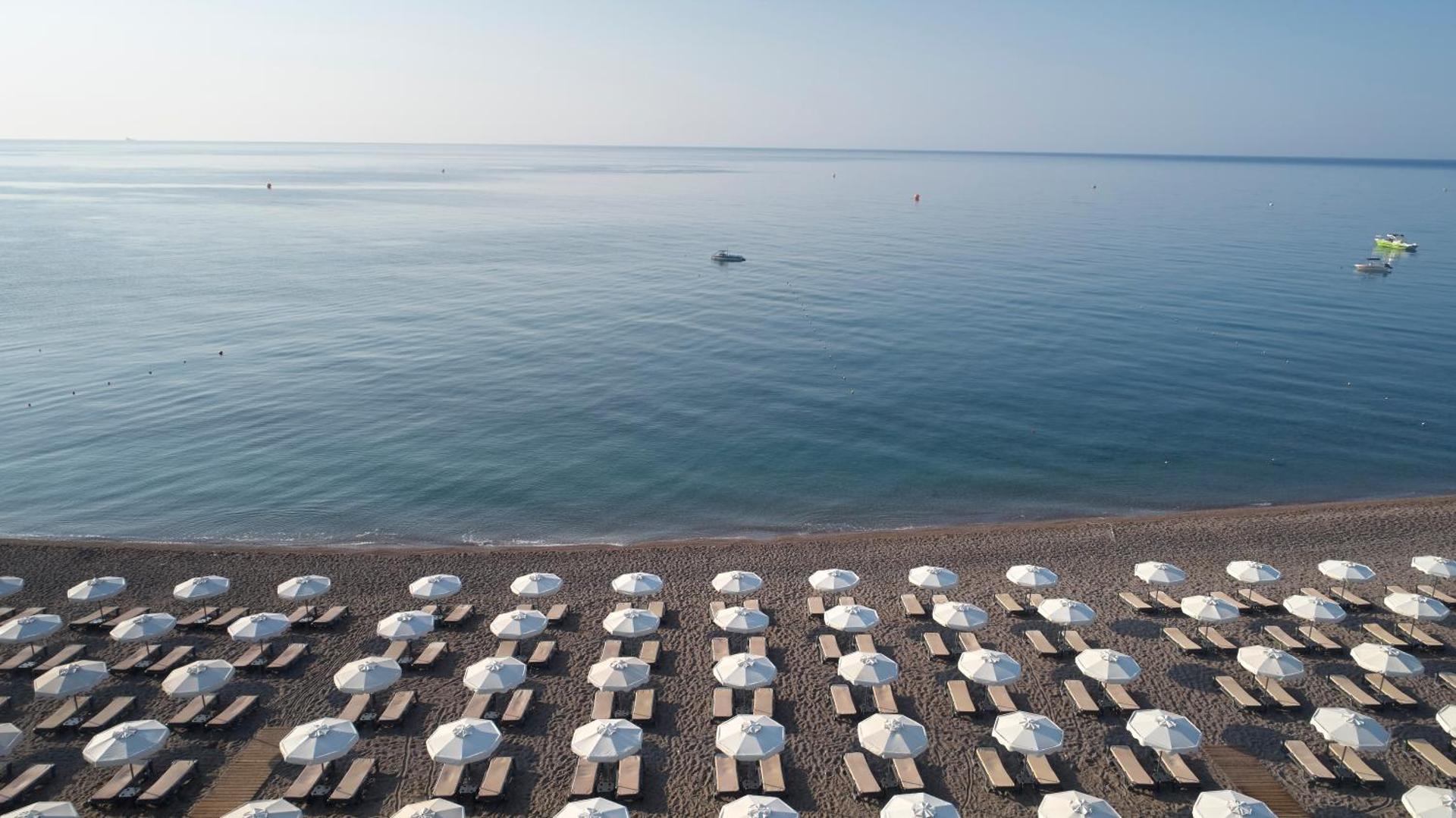 Skats uz Princess Andriana Resort & Spa pludmali no augšas — simetriskas saulessargu rindas un mierīga jūra rīta gaismā