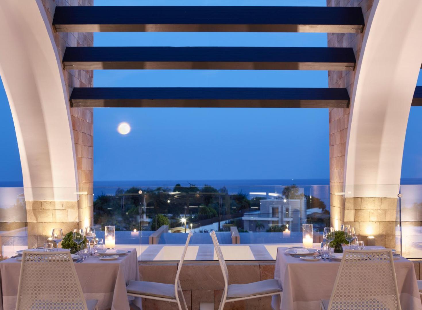 Princess Andriana Resort & Spa restorāna terase vakarā ar skatu uz jūru un mēnesi — eleganti klāti galdi divām personām