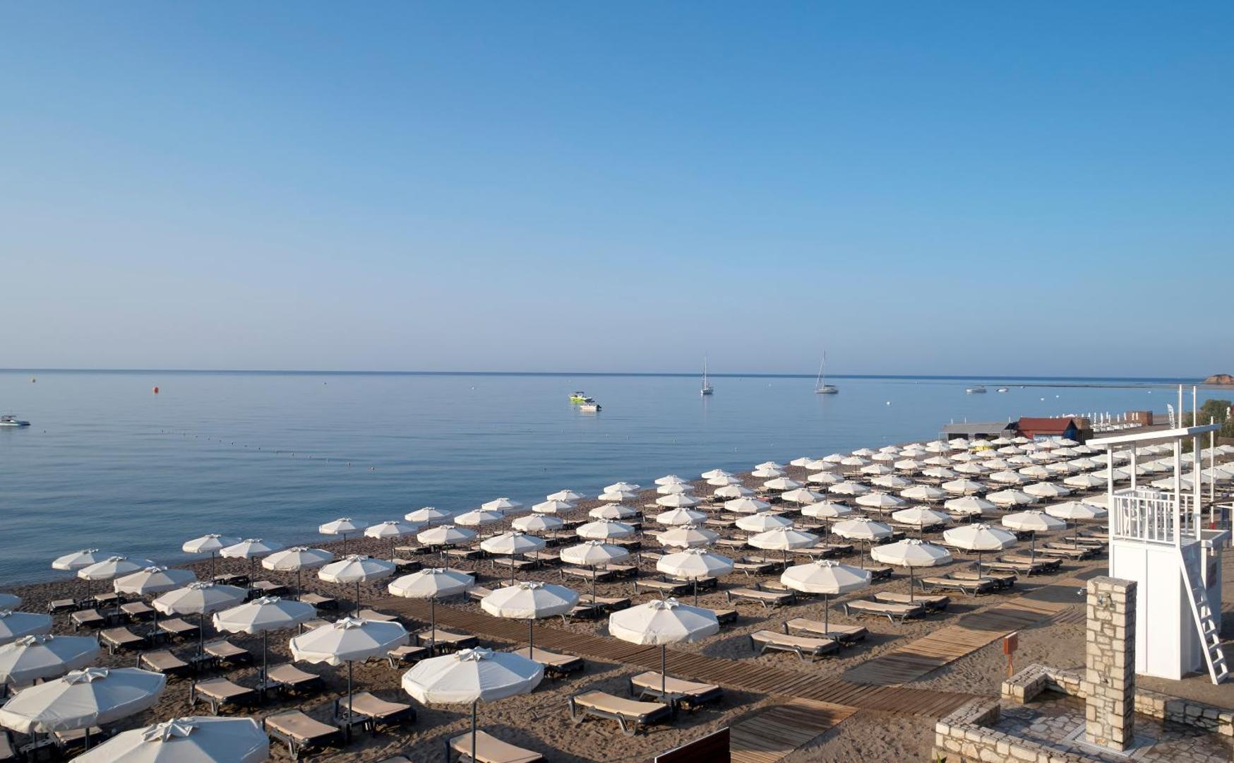 Princess Andriana Resort & Spa pludmales zona ar daudziem saulessargiem un guļamkrēsliem, fonā redzamas jahtas jūrā