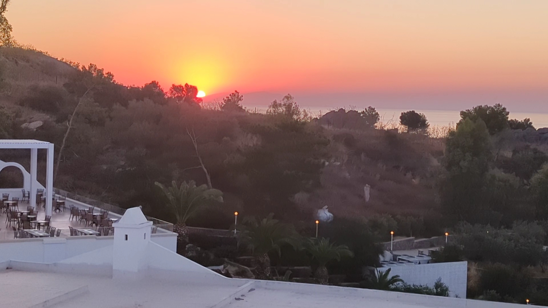 Saulriets virs Lindos Village Resort & Spa teritorijas Rodā, saskatāma āra terase ar galdiņiem un Vidusjūras ainava vakara gaismā