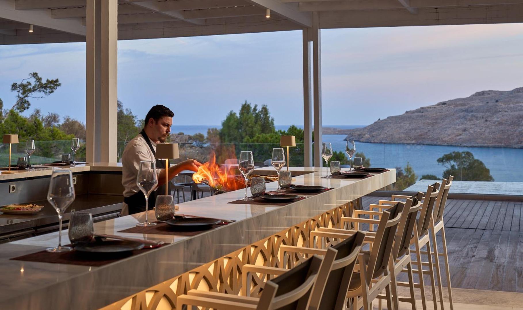 Lindos Village Resort & Spa ēdienkartes restorāns ar atvērto virtuvi, kur pavārs gatavo ēdienu uz uguns, fonā skats uz jūru