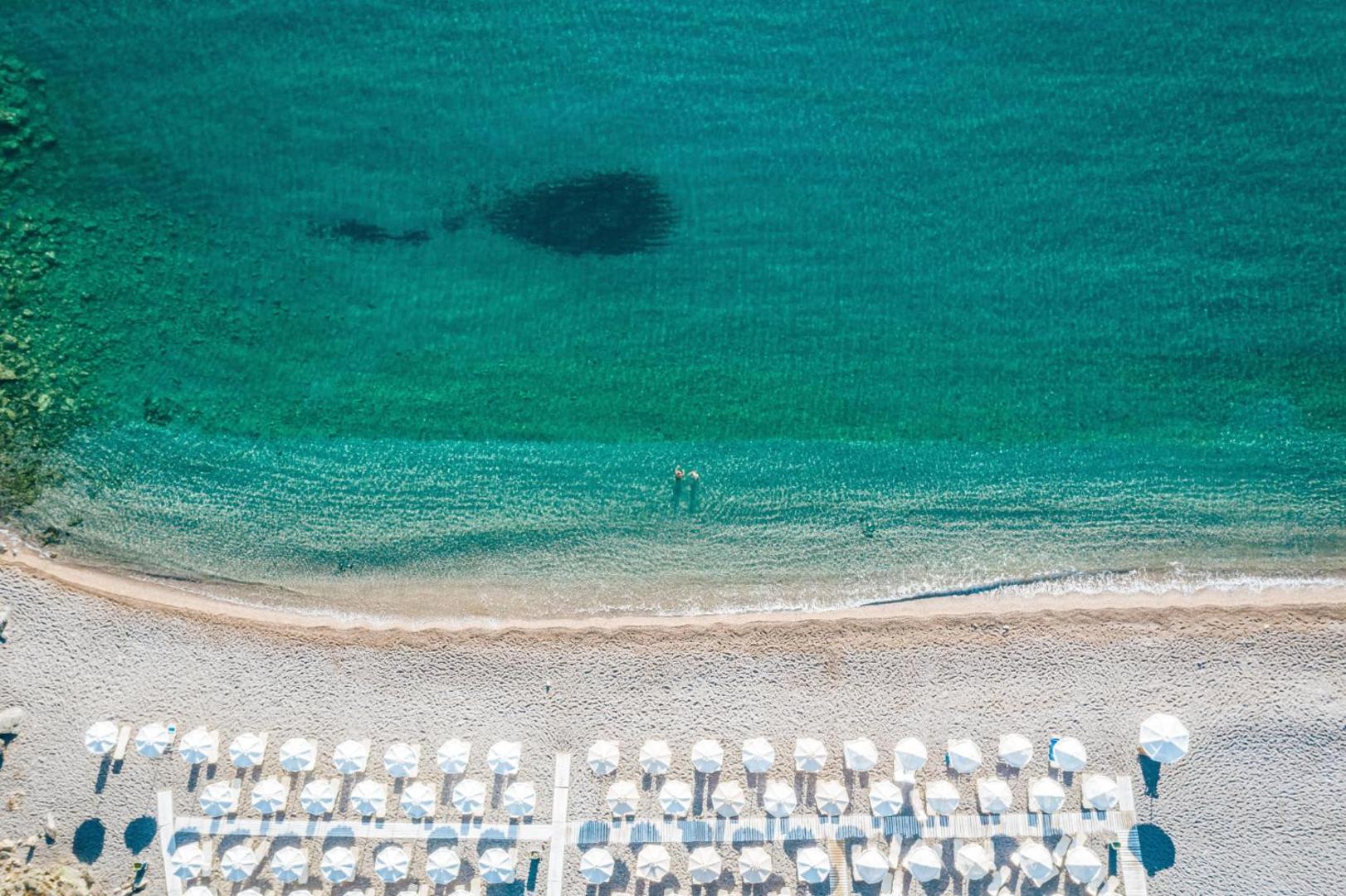 Aerofoto uzņēmums no Lindos Village Resort & Spa pludmales ar tirkīzzilu ūdeni un baltiem saulessargiem