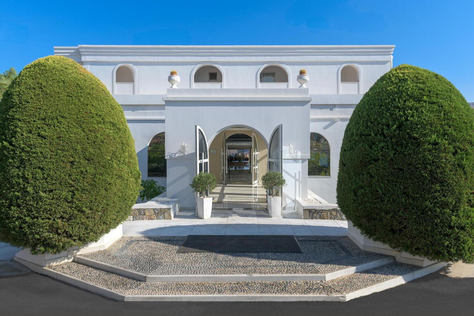 Lindos Village Resort & Spa galvenā ieeja ar baltu fasādi, arkveida durvīm un dekoratīviem apstādījumiem saulainā dienā