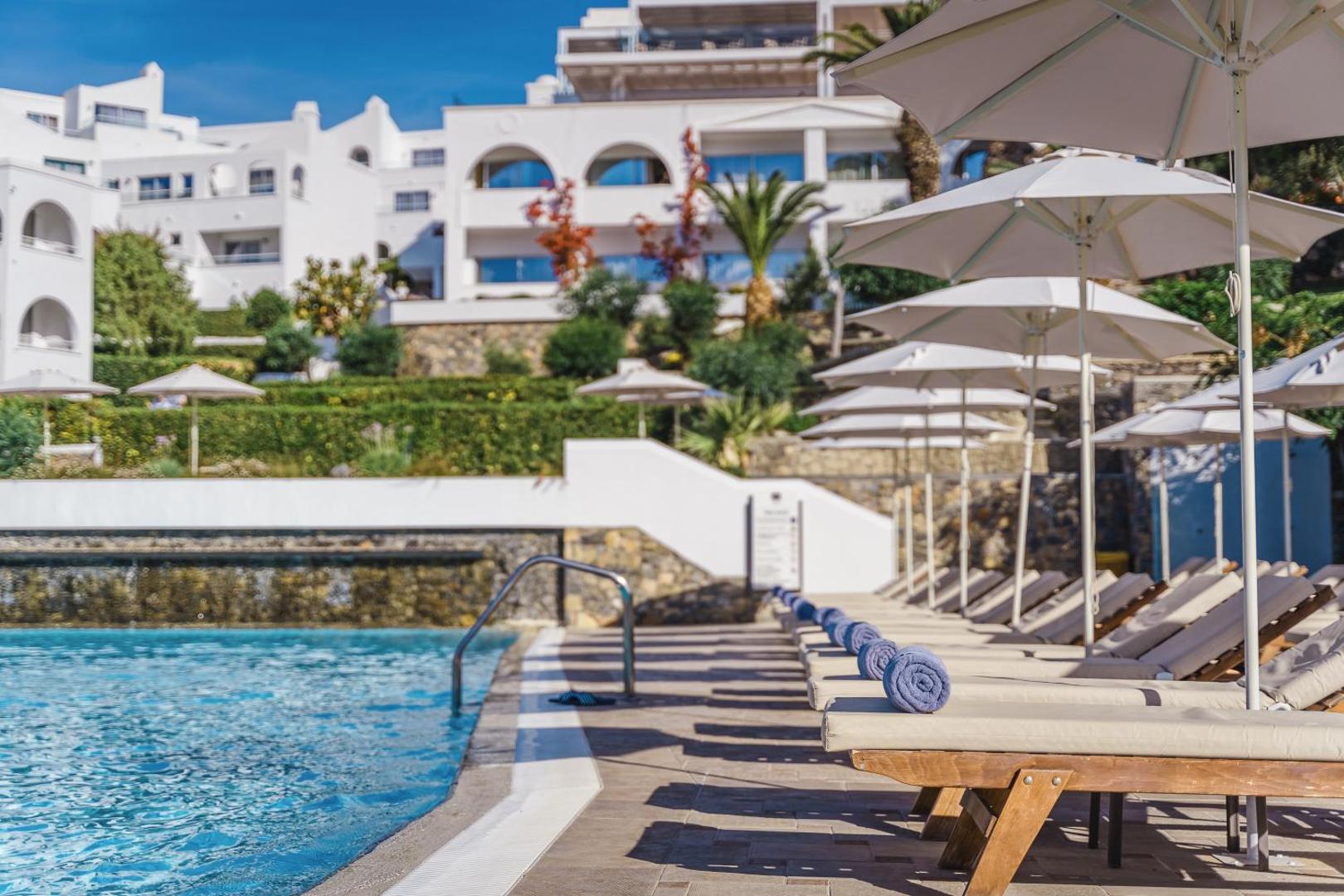Lindos Village Resort & Spa baseina zona ar koka sauļošanās krēsliem, ziliem dvieļiem un viesnīcas balto arhitektūru Rodas piekrastē