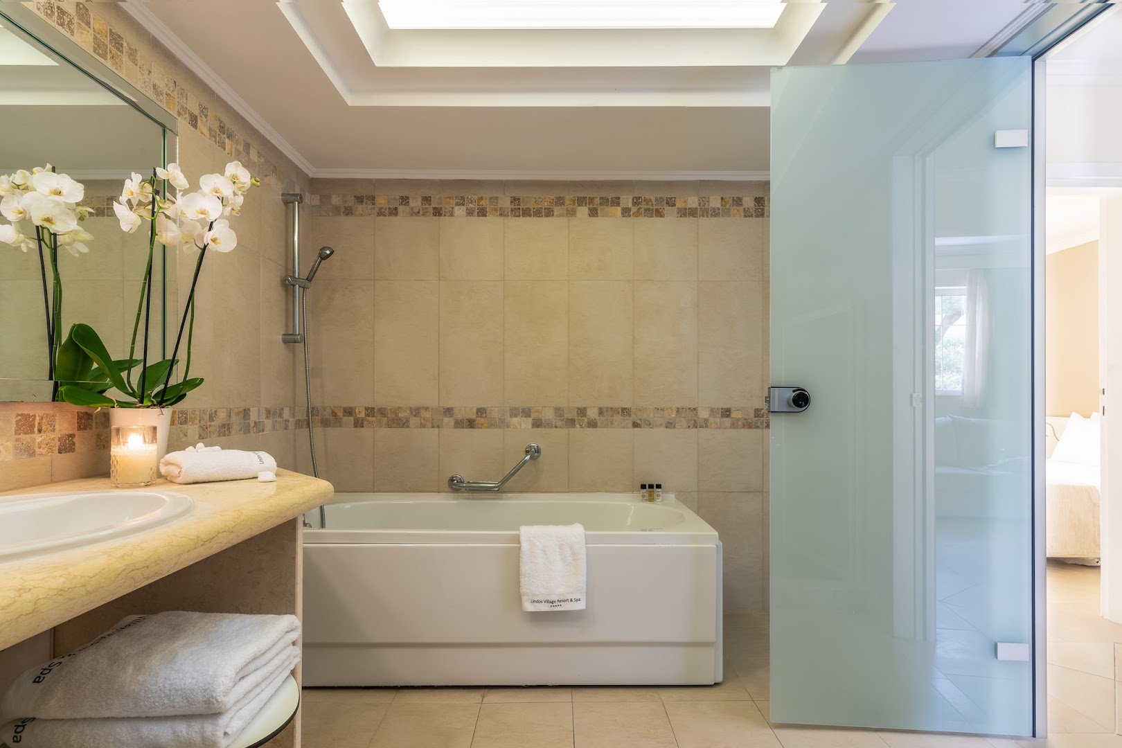 Lindos Village Resort & Spa vannas istaba ar gaišām flīzēm, vannu un orhidejām — elegants numura interjers Rodas salā
