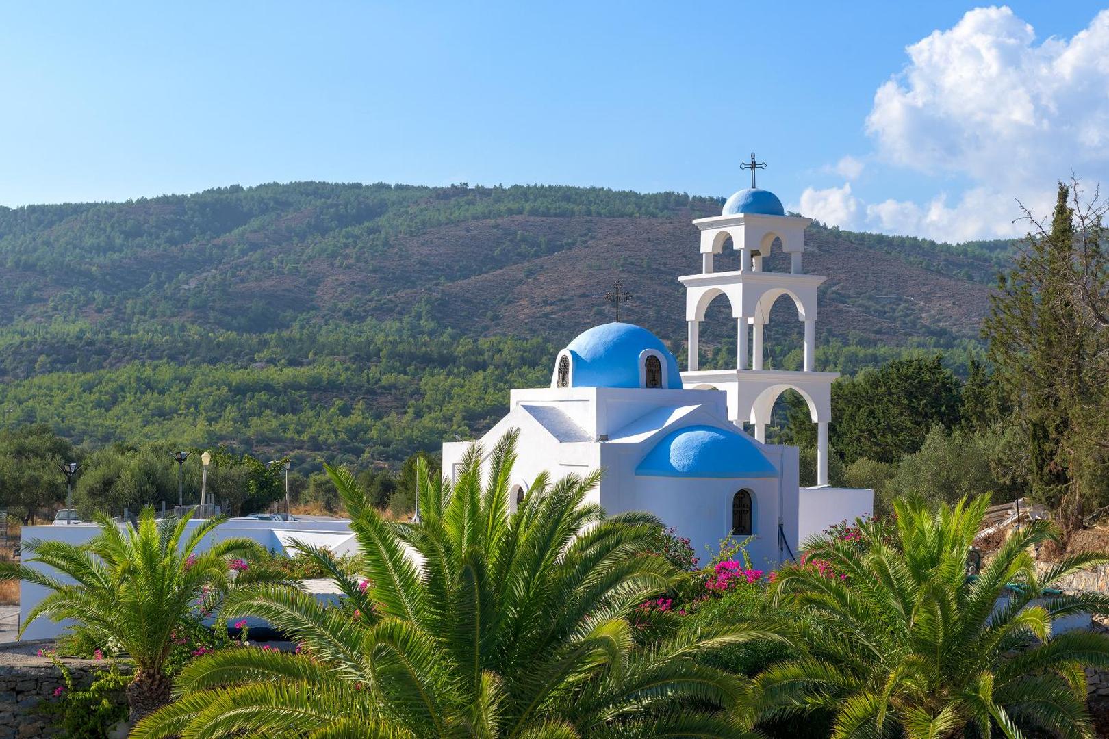 Tradicionāla grieķu baznīca ar ziliem kupoliem pie Lindos Village Resort & Spa Rodas salā — skats uz kalniem un palmām