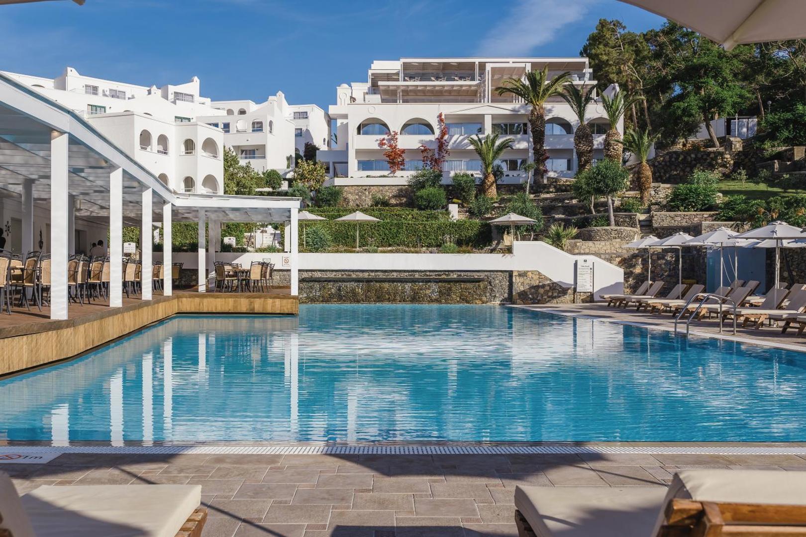 Skats uz Lindos Village Resort & Spa baseinu un restorāna terasi uz viesnīcas balto ēku fona Rodas salā