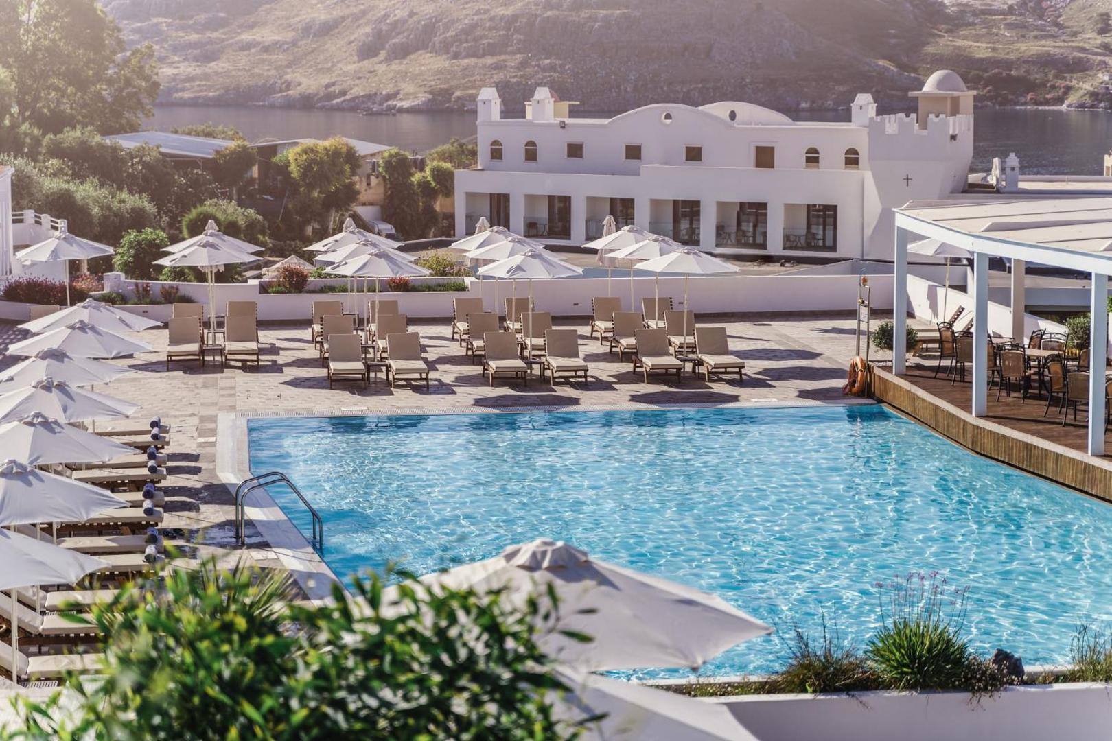 Lindos Village Resort & Spa plašā baseina terase ar skatu uz jūras līci un Rodas kalniem, aprīkota ar daudziem sauļošanās krēsliem