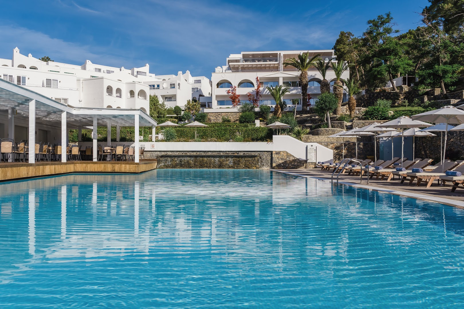 Lindos Village Resort & Spa galvenais āra baseins ar sauļošanās krēsliem un viesnīcas balto arhitektūru Rodas salā