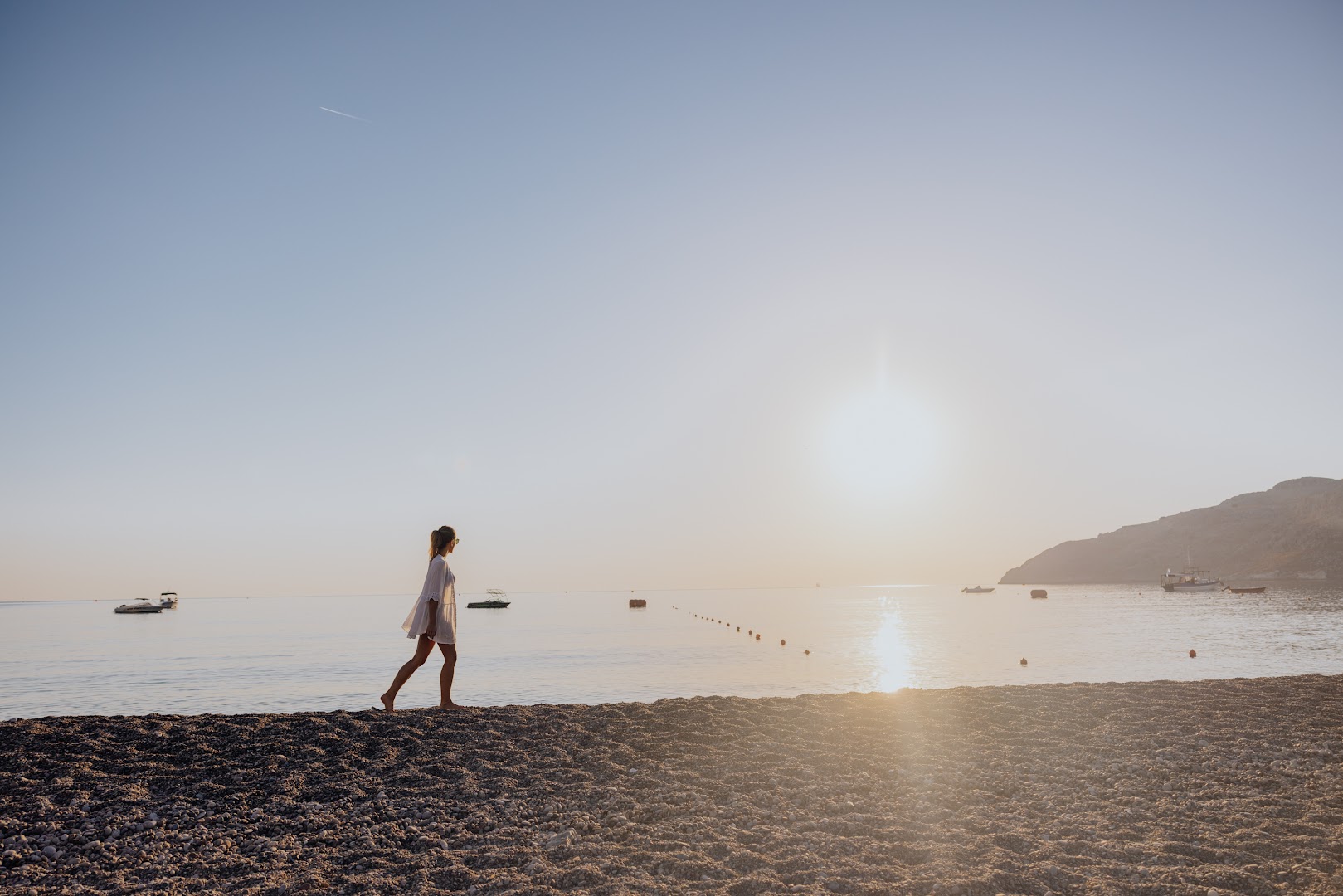 Lindos Village Resort & Spa oļu pludmale Rodas salā saulrieta laikā — sieviete pastaigājas gar jūras krastu pie kalniem