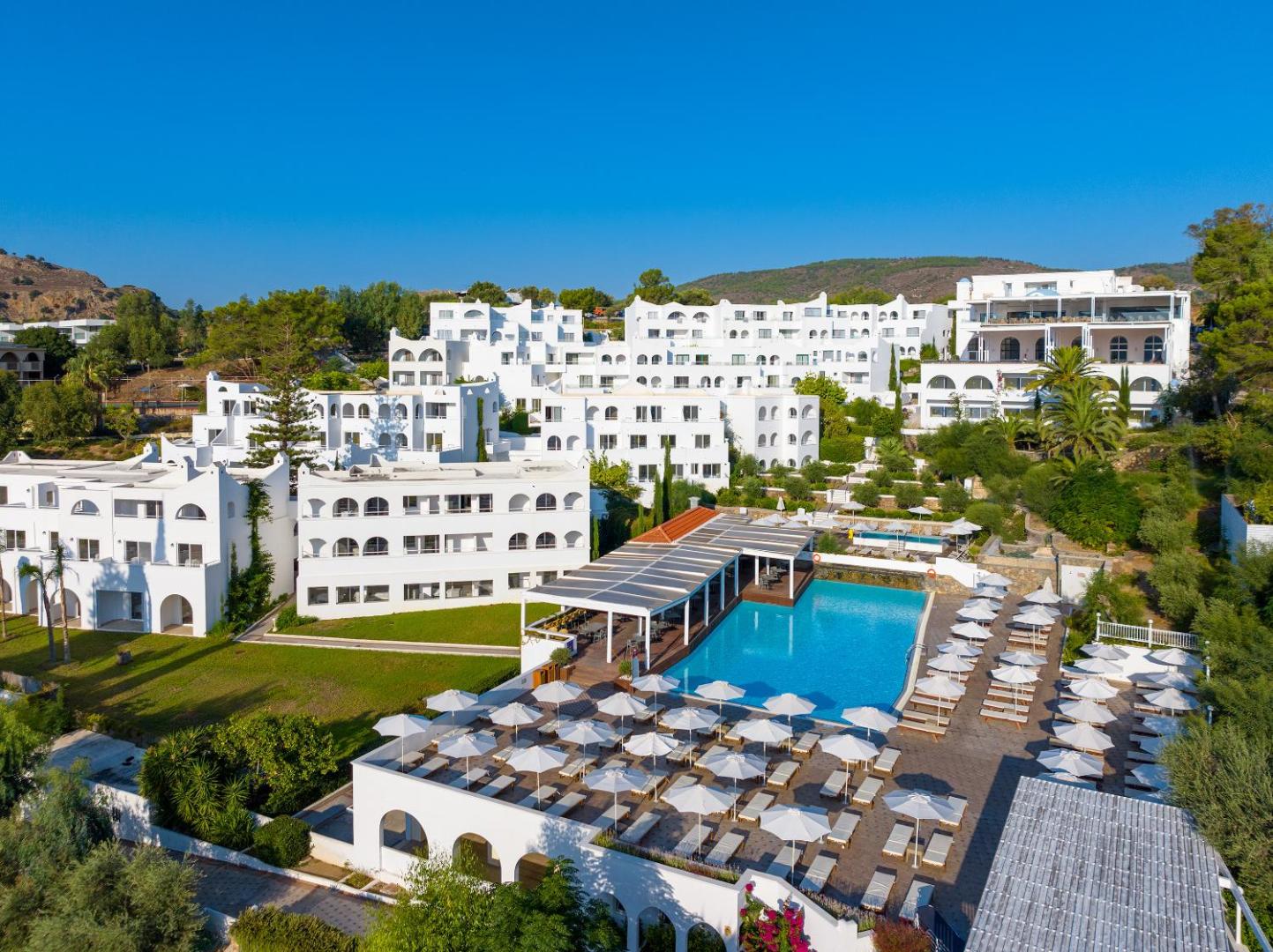 Lindos Village Resort & Spa galvenais āra baseins ar sauļošanās krēsliem, saulessargiem un baltām viesnīcas ēkām fonā Rodā